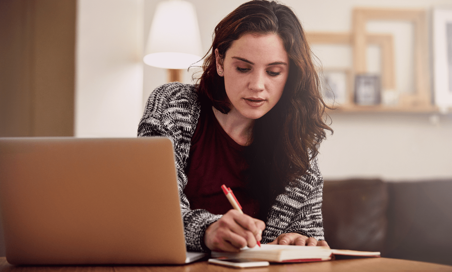 A young woman sits at a table in a cozy, softly lit room, journaling in a notebook with a red pen while looking thoughtfully at her notes. A laptop and phone rest nearby, suggesting a moment of reflection or goal-setting.