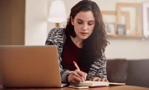A young woman sits at a table in a cozy, softly lit room, journaling in a notebook with a red pen while looking thoughtfully at her notes. A laptop and phone rest nearby, suggesting a moment of reflection or goal-setting.