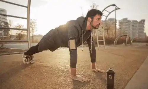 A man in athletic wear holds a focused plank position outdoors at sunrise, wearing earbuds and using a smartphone armband, with a water bottle nearby—symbolizing discipline, movement, and progress.