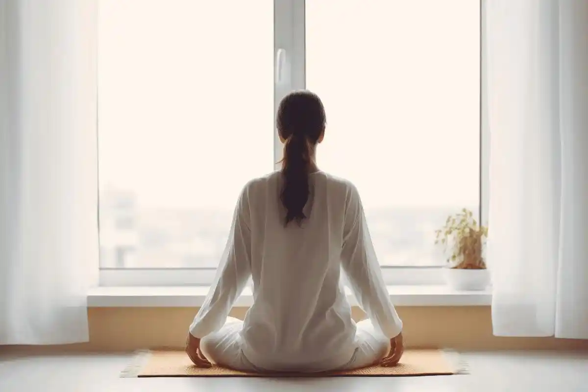 Rear view of a person in white loungewear meditating cross-legged on a yoga mat in front of a large window. The setting is serene, with soft daylight and minimal decor.