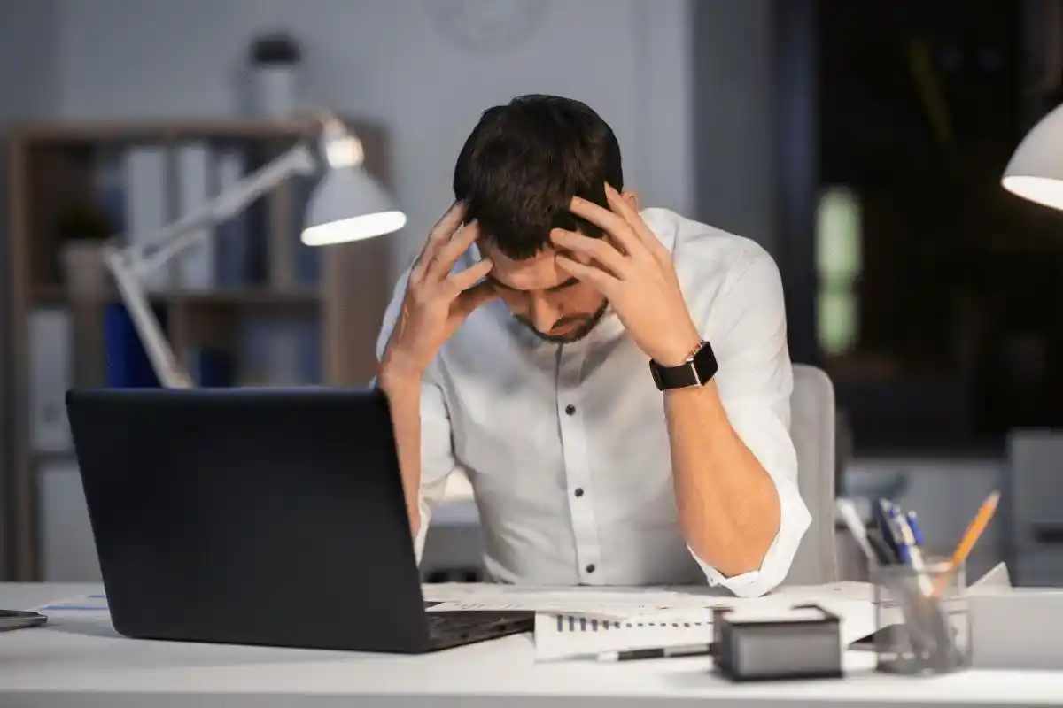 Frustrated man sitting at a desk in a dimly lit office, holding his head in his hands. He appears overwhelmed by work, surrounded by a laptop and paperwork.