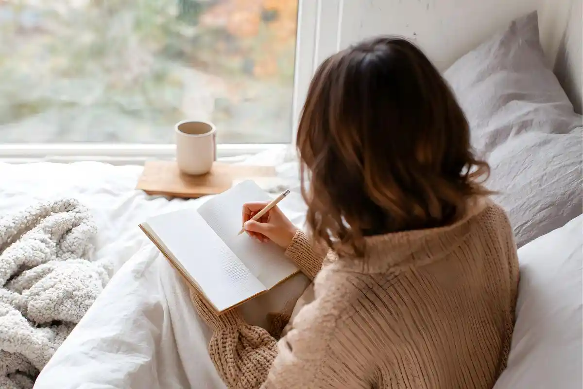 Person journaling in bed beside a window, wrapped in a blanket with a warm drink nearby—reflecting a quiet, self-care moment."